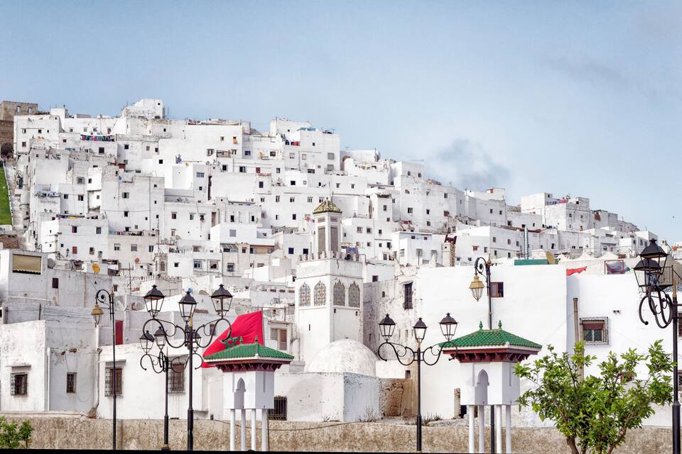 Un pueblo en una ladera con casas blancas, una destacada bandera roja, farolas tradicionales y una mezquita con cúpula verde.