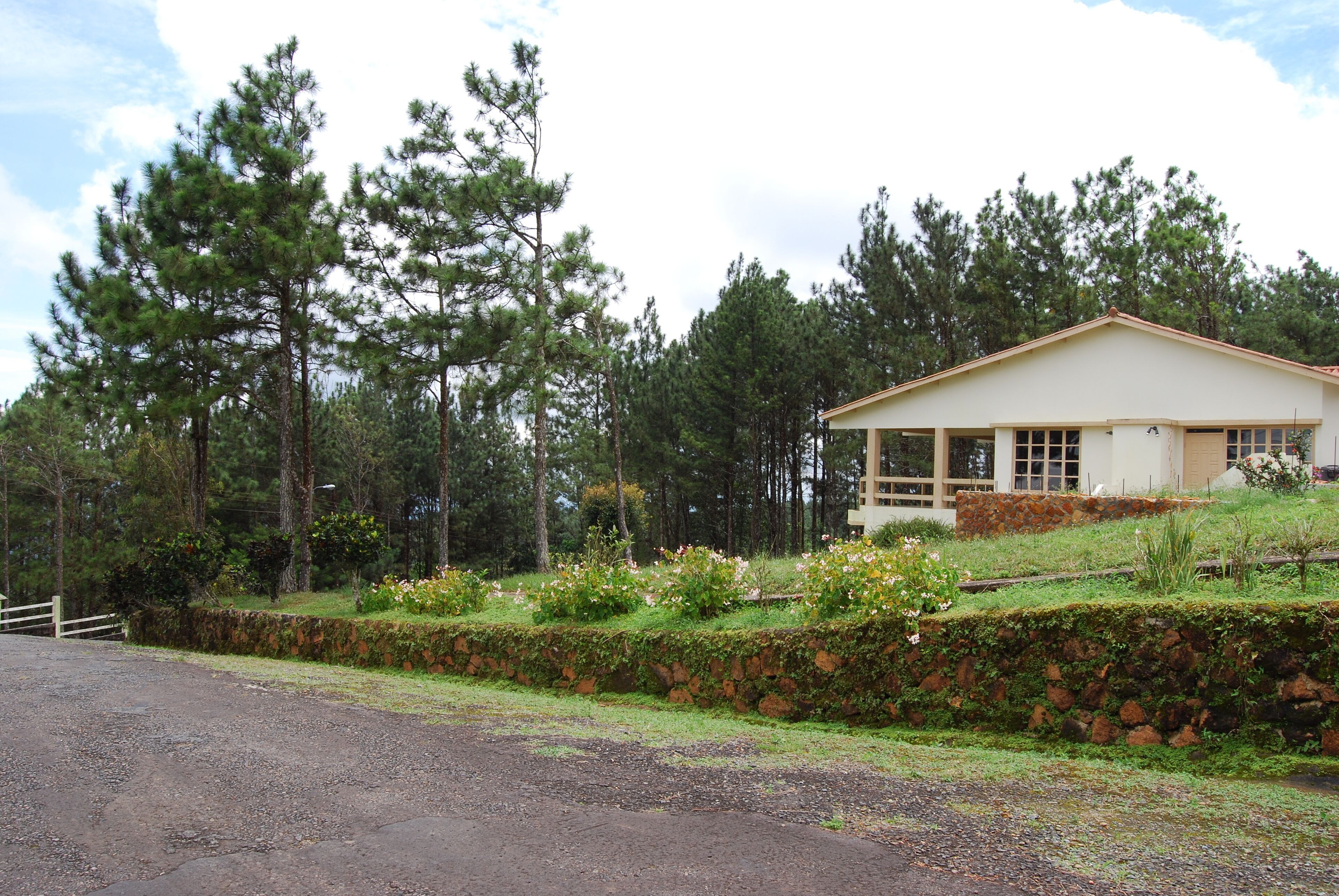 A house with a sloped roof, surrounded by a stone wall and a grassy area with flowers.