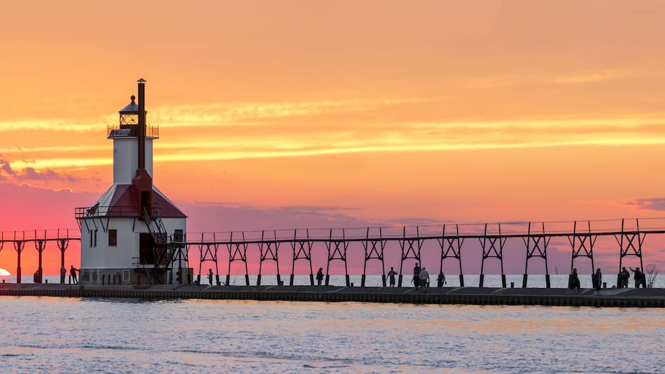 A coastal pier with a lighthouse and a white building during sunset.