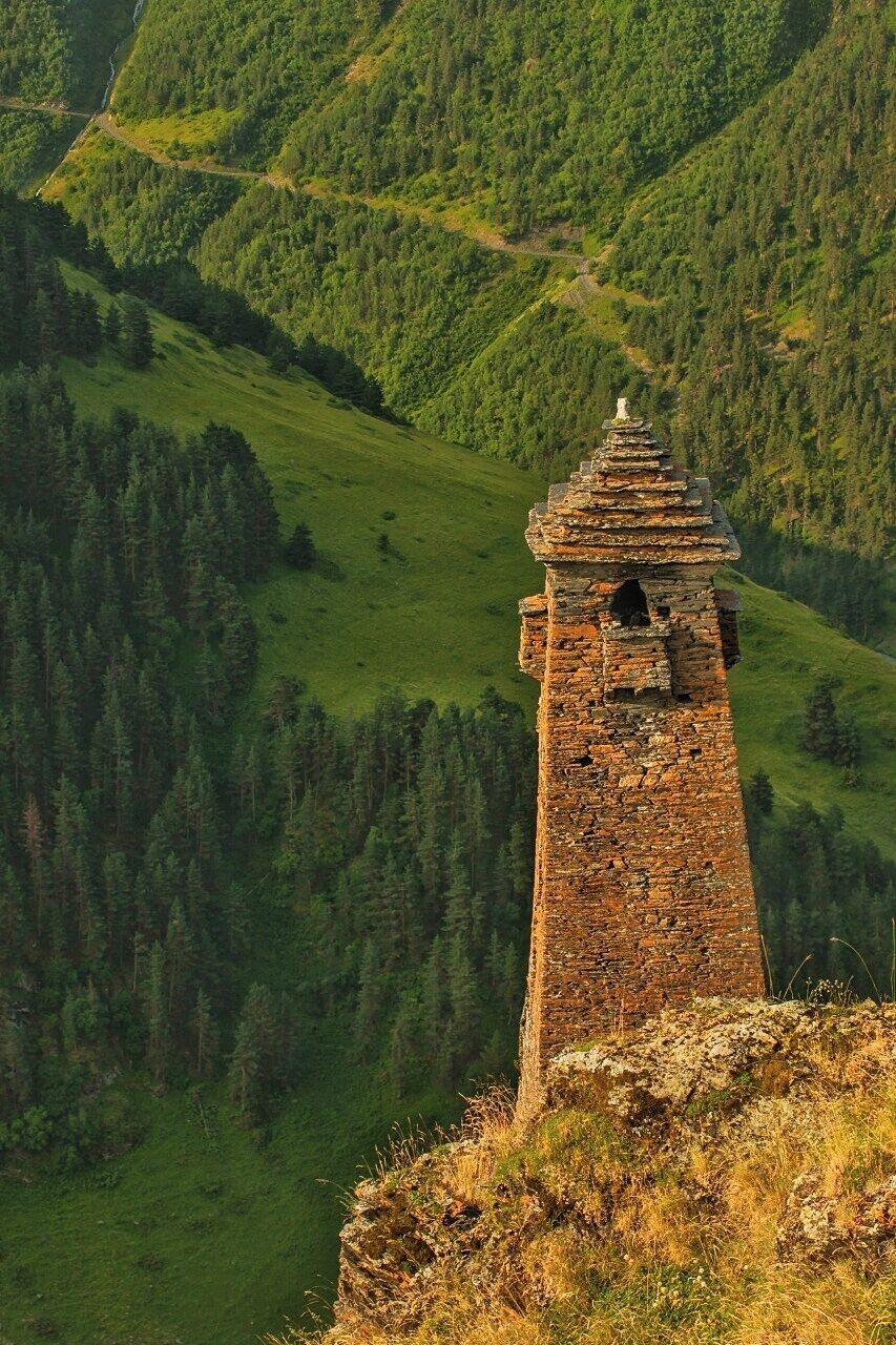 Ein steinerner Turm auf einer Hügelsicht mit einem bewaldeten Tal im Hintergrund.