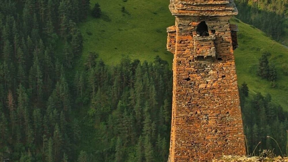 Une tour de pierre, située sur le versant d’une colline, avec une vallée boisée en arrière-plan.