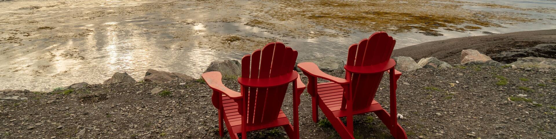A pair of adirondack chairs at the edge of the beach overlooking the ocean, clouds reflecting in the still bay water in front