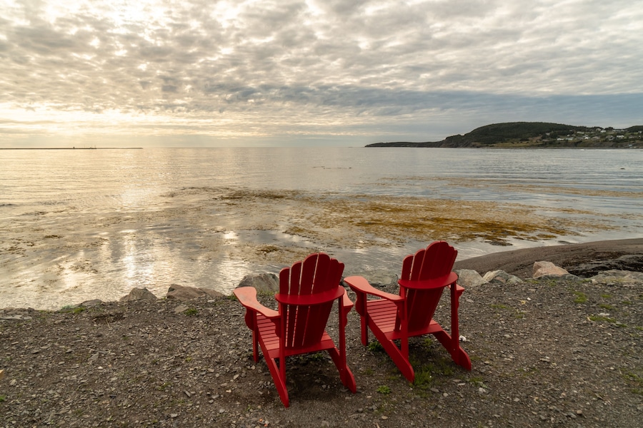 A pair of adirondack chairs at the edge of the beach overlooking the ocean, clouds reflecting in the still bay water in front
