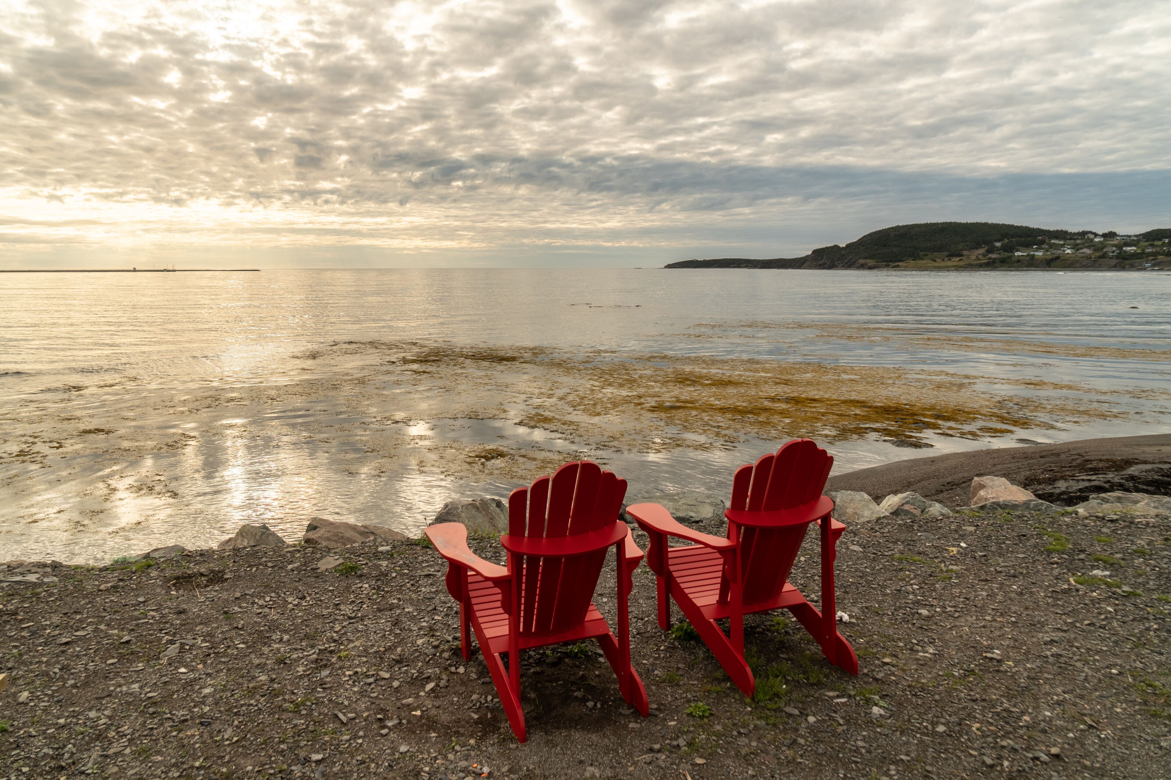 Two red Adirondack chairs facing a calm lake with a distant shoreline and cloudy sky.