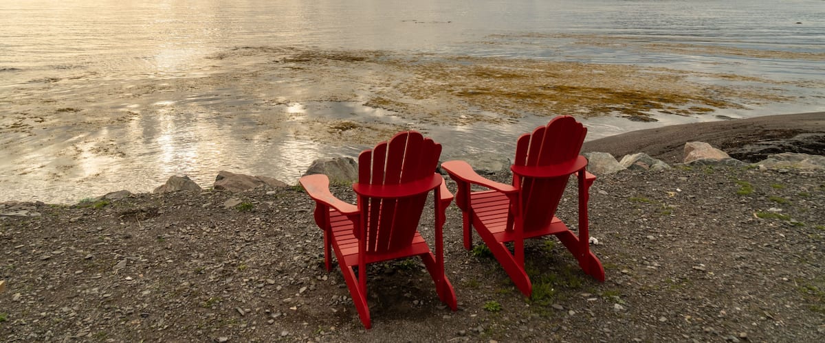 Two red Adirondack chairs facing a calm lake with a distant shoreline and cloudy sky.