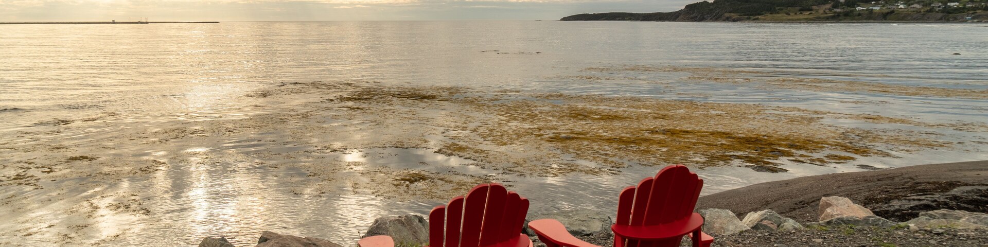 Two red Adirondack chairs facing a calm lake with a distant shoreline and cloudy sky.