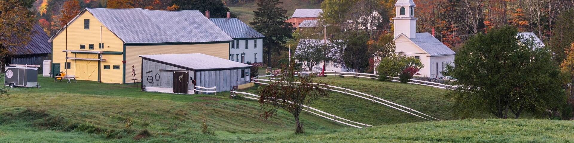 Paisaje rural con una casa amarilla, una iglesia blanca y una colina arbolada con follaje otoñal.