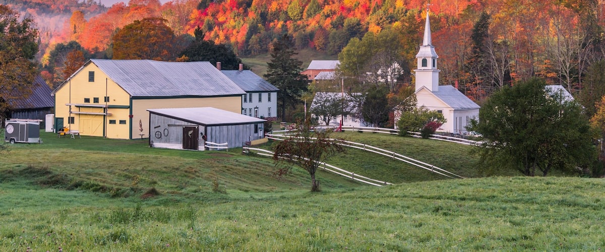 A rural landscape with a yellow house, a white church, and a forested hill with autumn foliage.