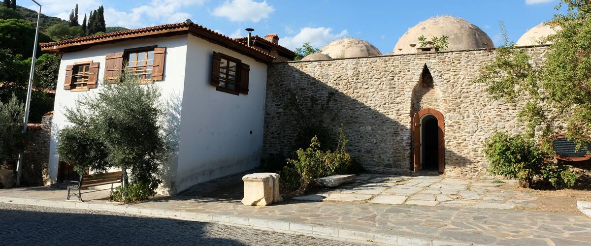 A traditional building with stone walls and wooden shutters, a cobblestone street, and domed structures in the background.