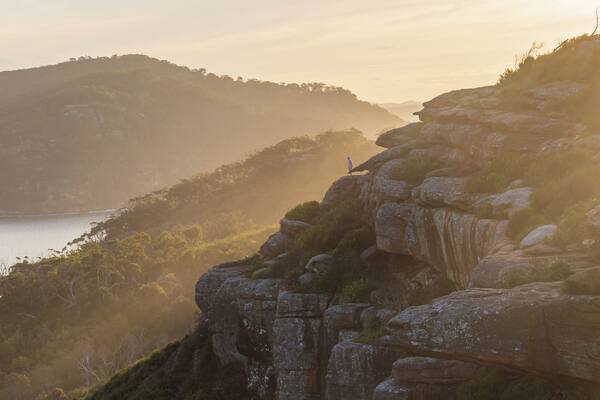 A person standing on a rocky outcrop overlooking a body of water and hills.