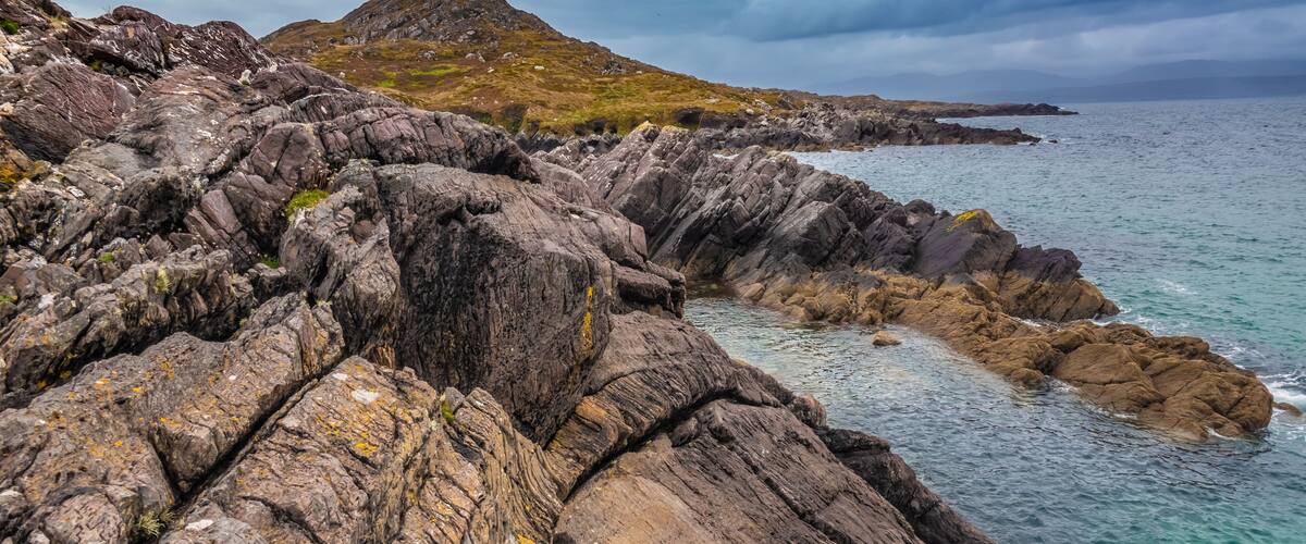 Rugged coastal rocks, a distant mountain, and a calm sea under cloudy skies.