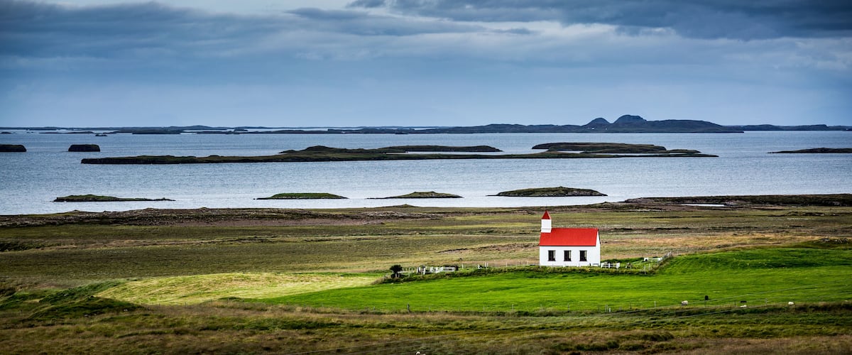 A coastal landscape with a small white house with a red roof, green fields, and rocky outcrops in the water.