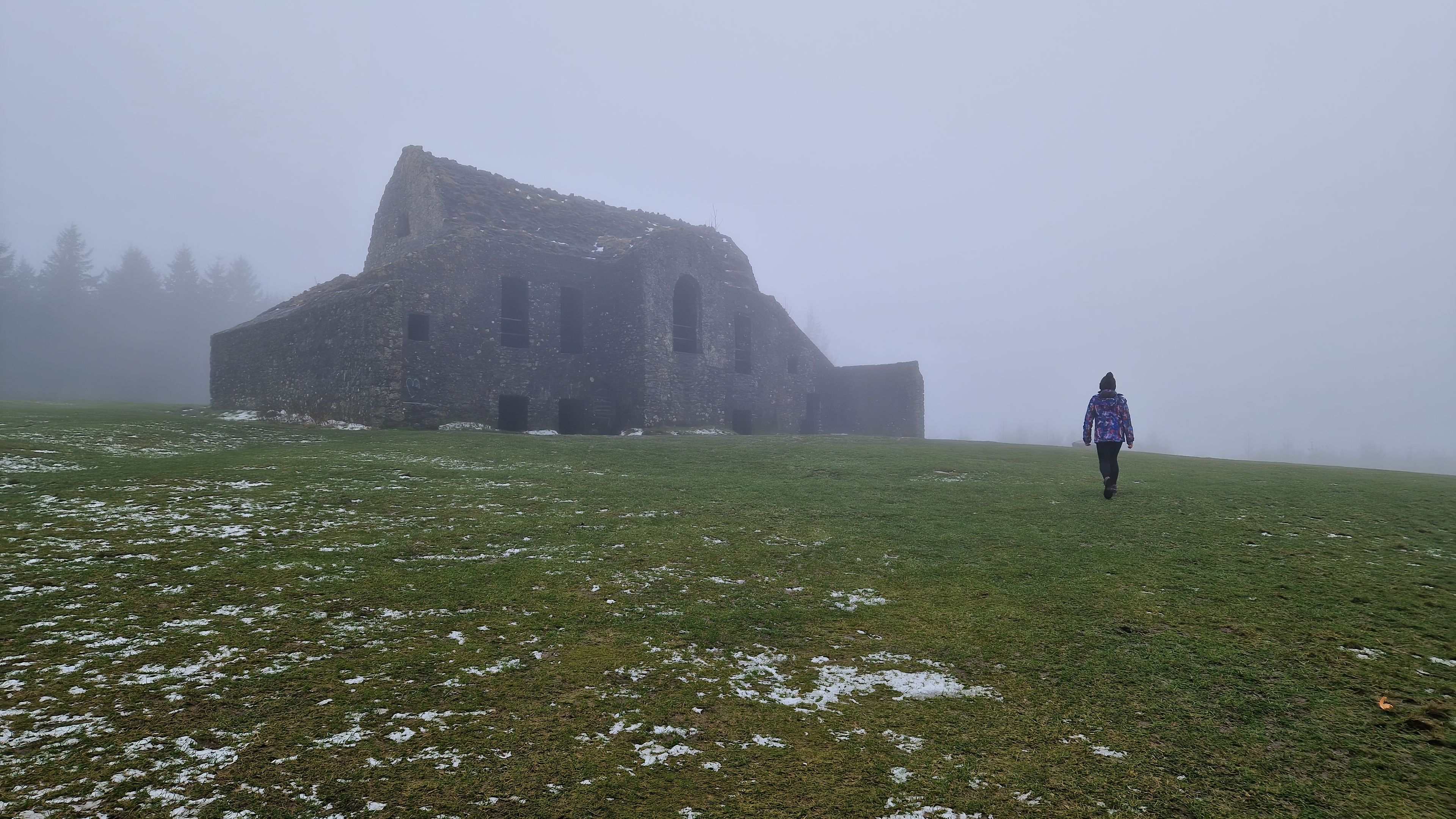 A person walking towards a large, dilapidated stone building in a foggy, grassy field.