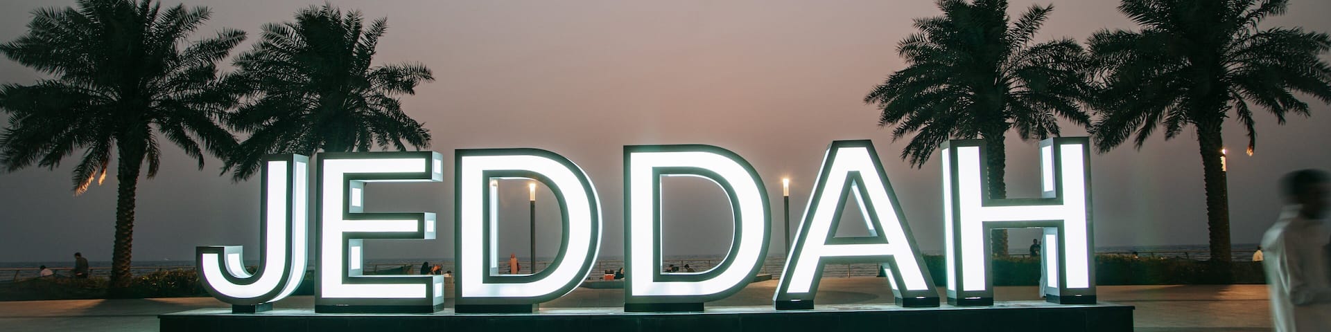 Illuminated letters spelling "JEDDAH" on a platform with palm trees in the background.