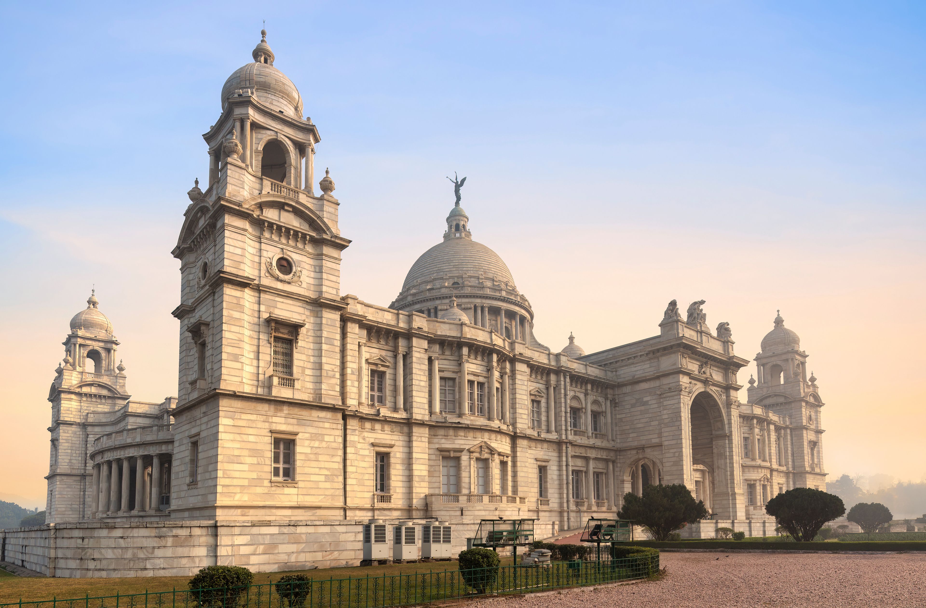 A grand colonial-style building with multiple domes and towers, surrounded by a well-maintained garden and a gravel pathway.