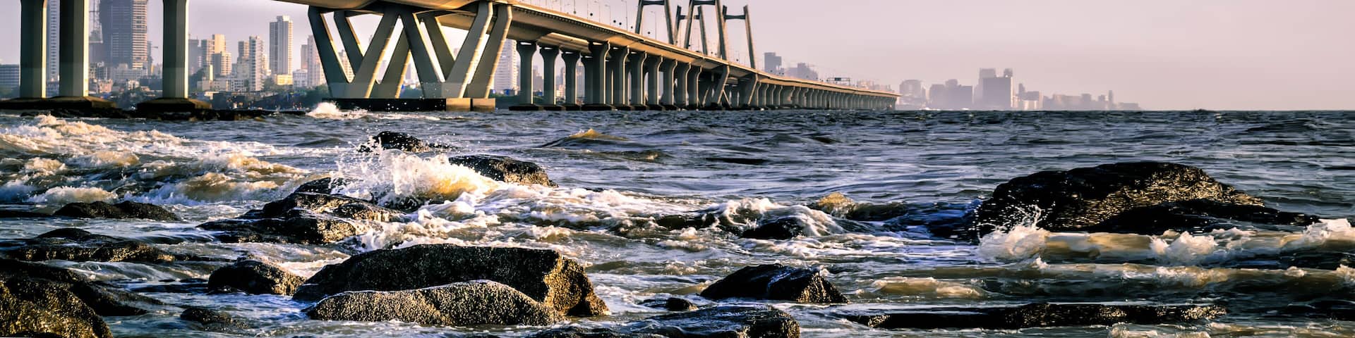 A suspension bridge over a body of water with waves crashing against rocks in the foreground.