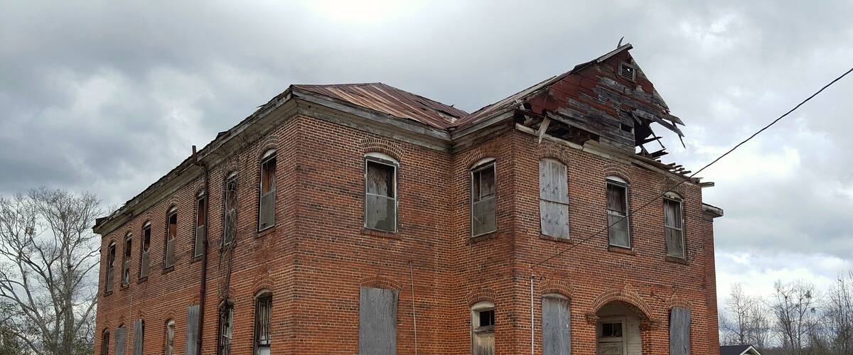 A large, two-story brick building with a damaged roof and boarded-up windows.