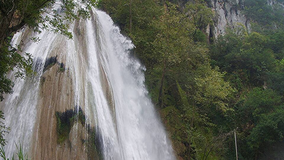 A waterfall cascading over a rocky cliff, surrounded by lush greenery and a bridge with a metal railing.