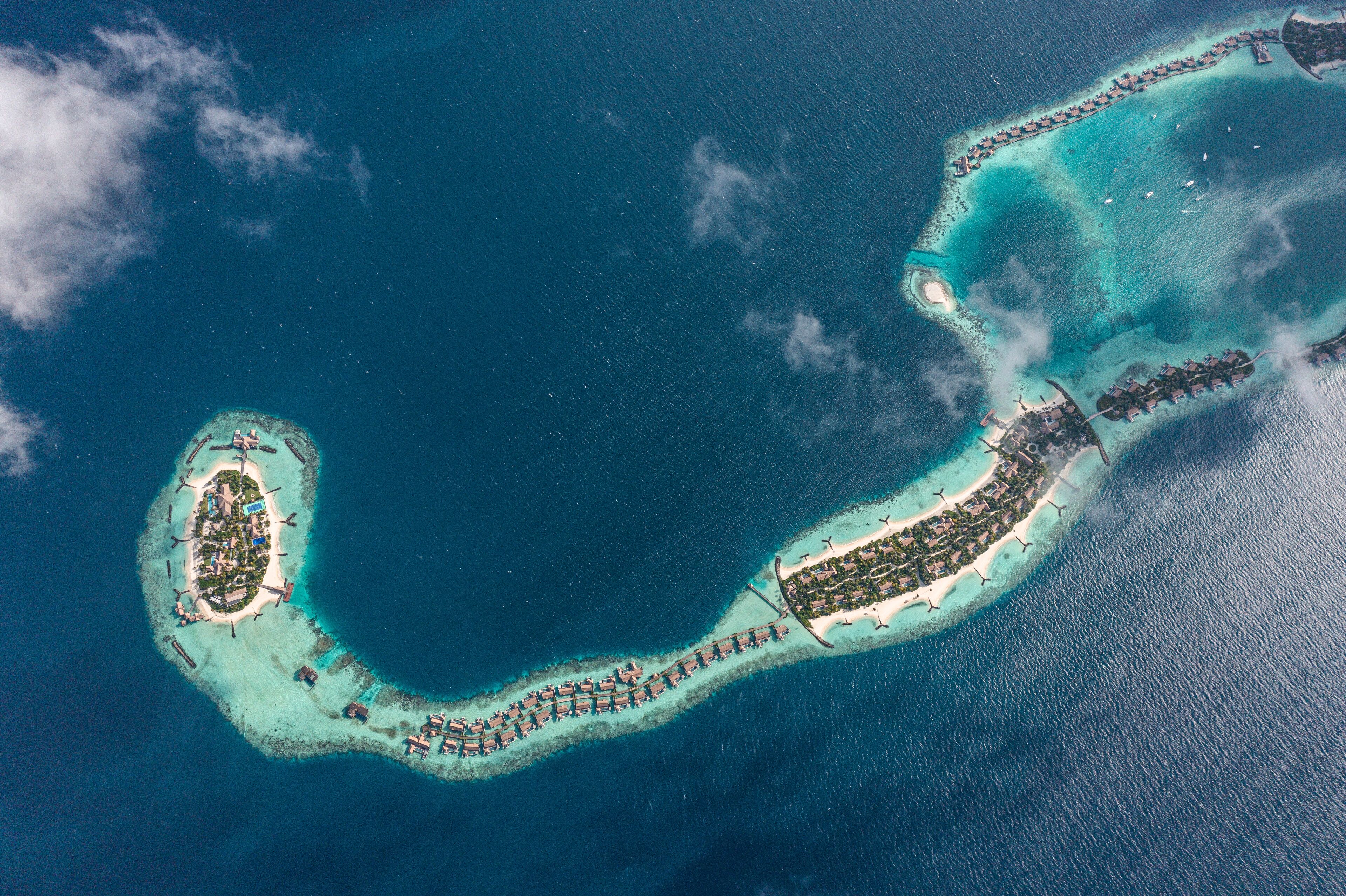An aerial view of a tropical island with clear turquoise waters, a long crescent-shaped resort, and a small central island with a lighthouse.