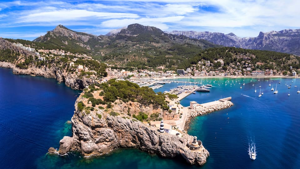 A coastal town with a marina, rocky cliffs, and mountains in the background.