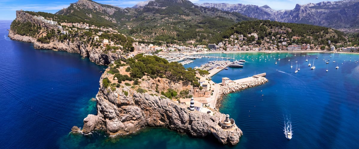 A coastal town with a marina, rocky cliffs, and mountains in the background.