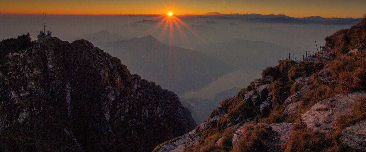 A mountain landscape at sunset with the sun visible above the horizon.