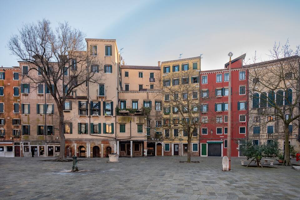 A historic square with multi-story buildings, a fountain, and benches.