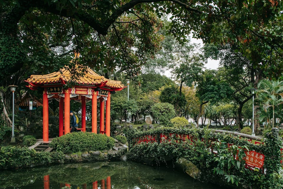 A traditional Chinese pavilion with red pillars and golden roof, surrounded by lush greenery and a pond.