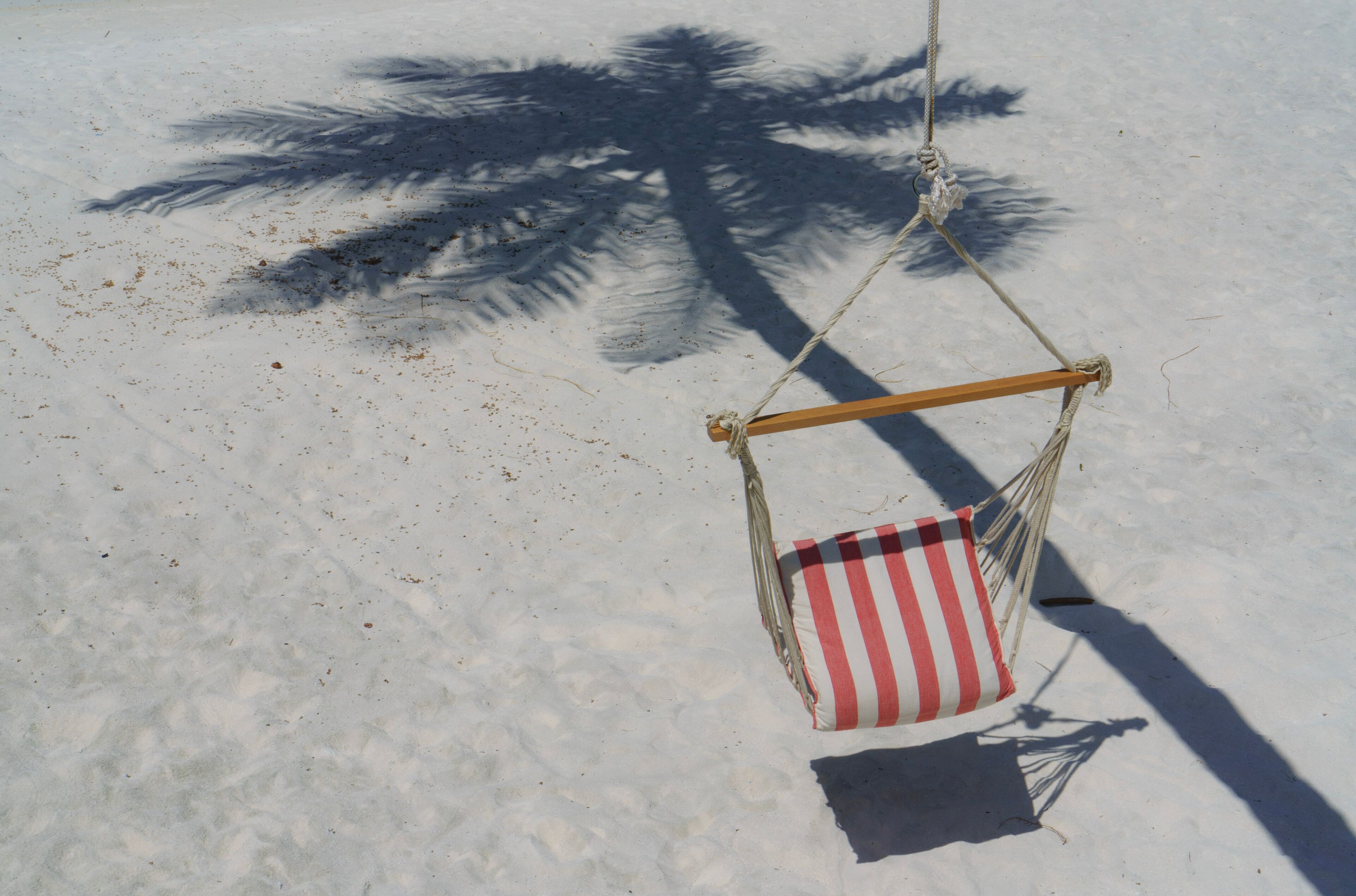 A beach hammock with a striped pillow, a palm tree shadow, and sand.