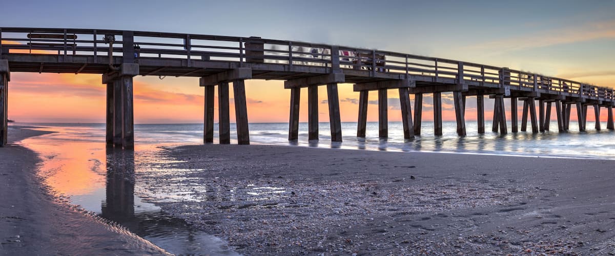 A pier extending into calm waters at sunset with a beach in the foreground.
