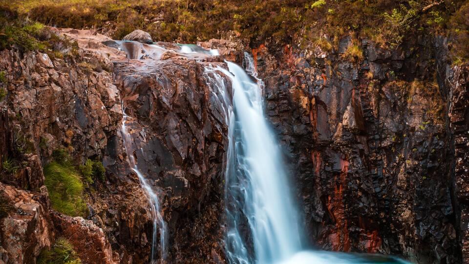 A waterfall cascading into a turquoise pool surrounded by rugged mountains and sparse vegetation.