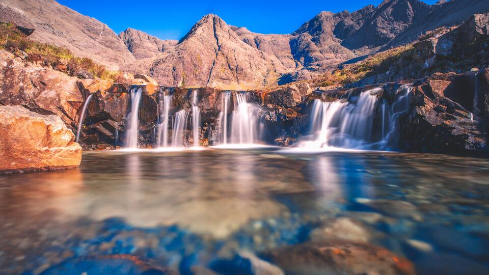 A waterfall cascading over rocks into a clear pool, surrounded by mountains.