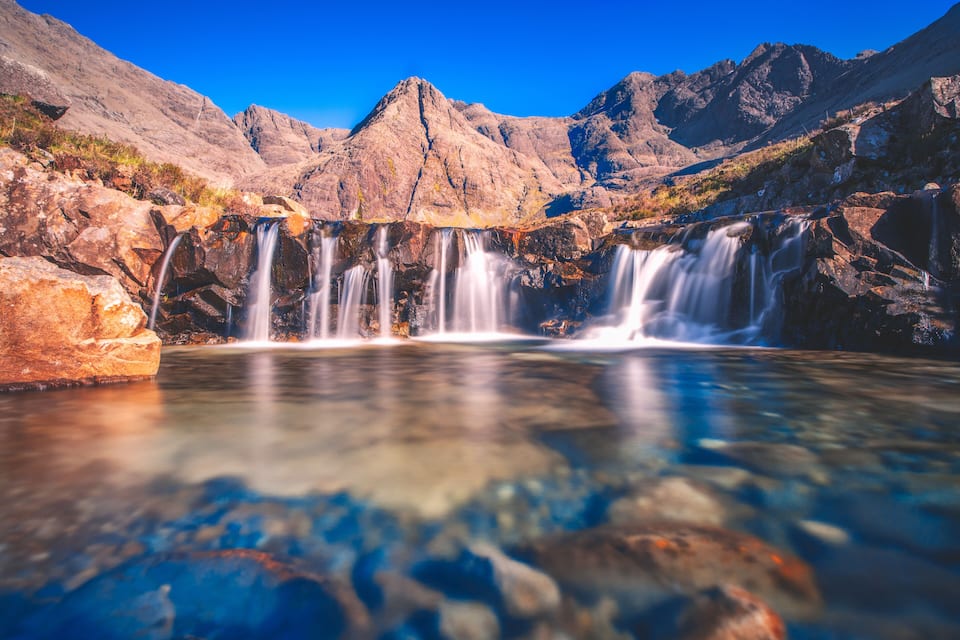A waterfall cascading over rocks into a clear pool, surrounded by mountains.