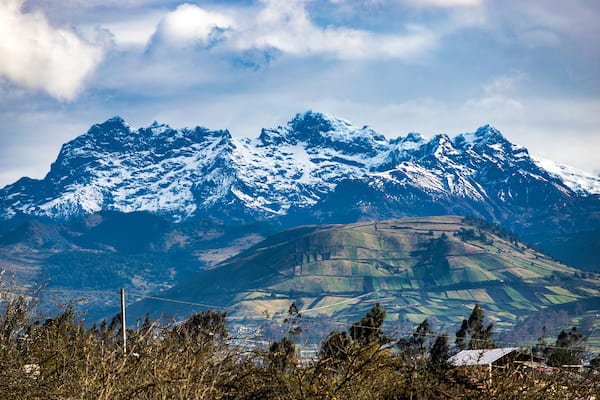 Snow-capped mountains, a valley with fields, and a distant house.