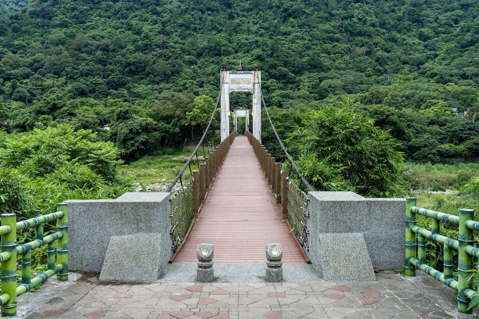 Un puente colgante de madera con barandillas de piedra, rodeado de un bosque verde y frondoso.