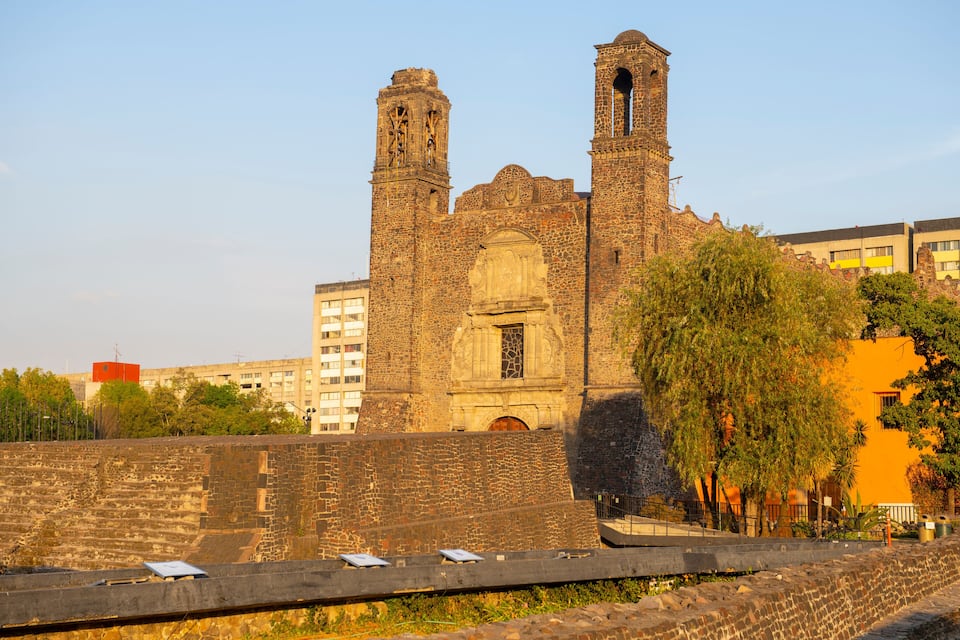 Uma igreja de pedra histórica com duas torres sineiras, cercada por edifícios modernos e árvores.