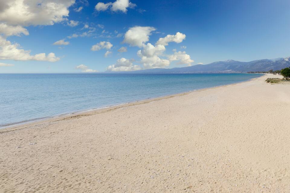 A sandy beach with clear blue water and distant mountains under a partly cloudy sky.