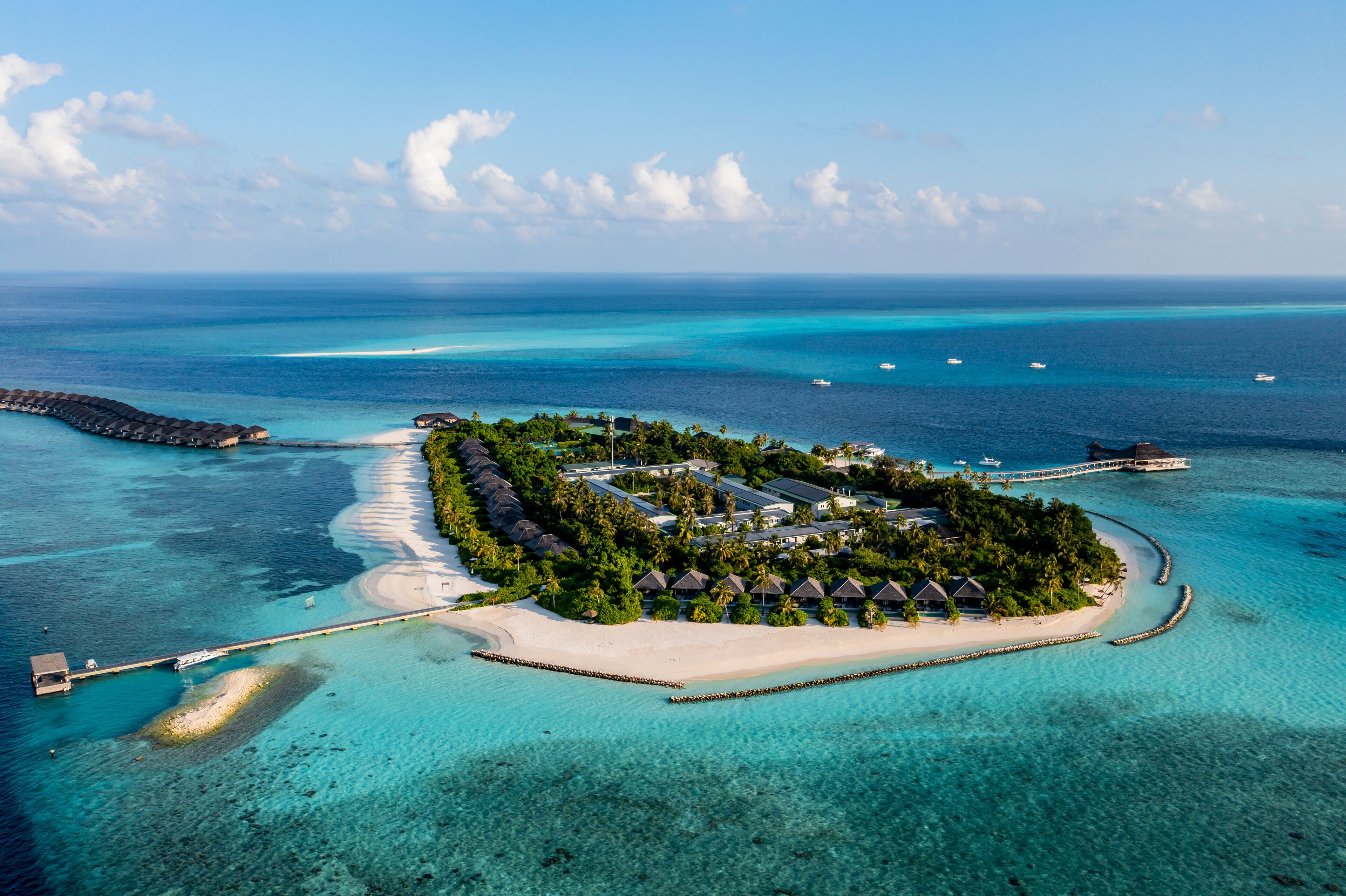 An aerial view of a tropical island resort with overwater bungalows, clear turquoise waters, and a white sandy beach.