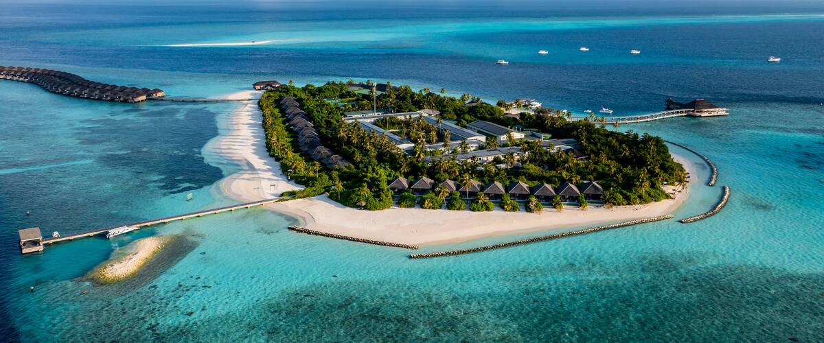 An aerial view of a tropical island resort with overwater bungalows, clear turquoise waters, and a white sandy beach.