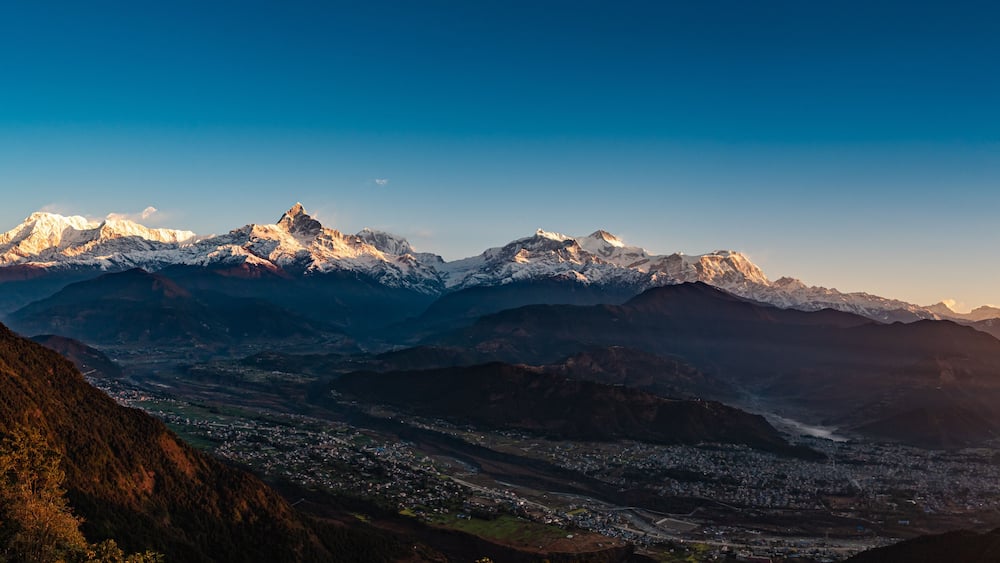 A mountain range with snow-capped peaks, a valley with a town, and a clear sky.