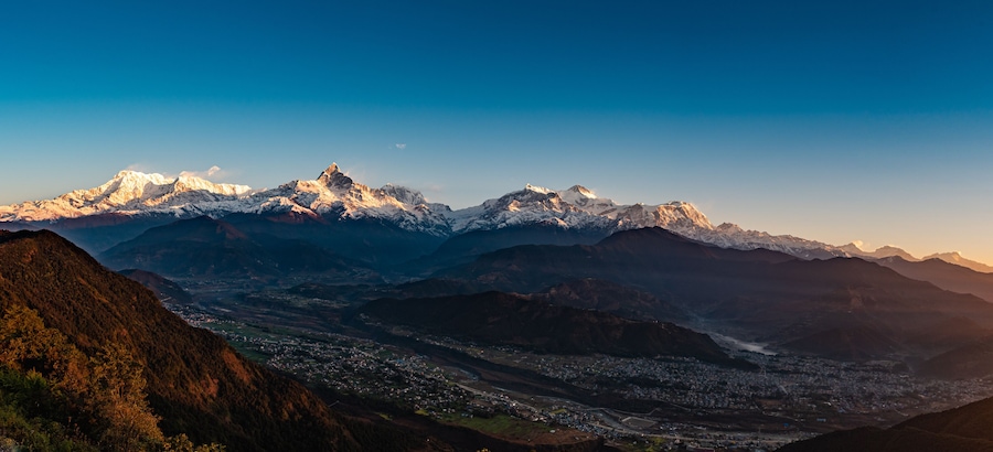 A mountain range with snow-capped peaks, a valley with a town, and a clear sky.
