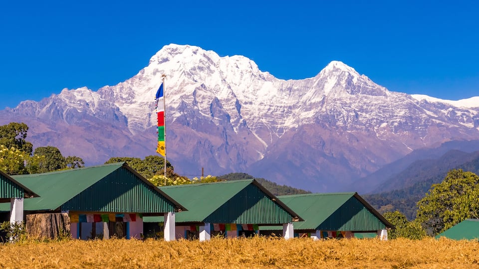A mountain range with snow-capped peaks, a flagpole with multiple flags, and green-roofed buildings in a field.