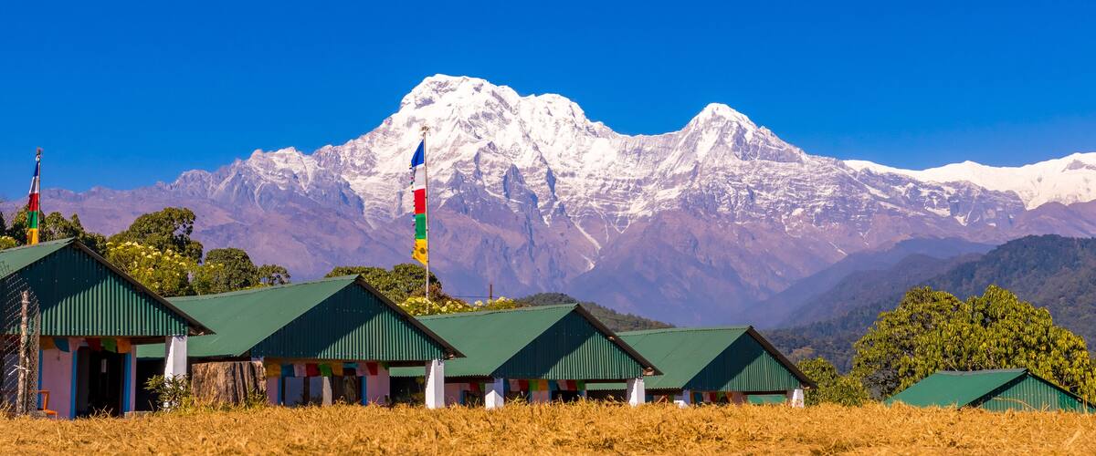A mountain range with snow-capped peaks, a flagpole with multiple flags, and green-roofed buildings in a field.
