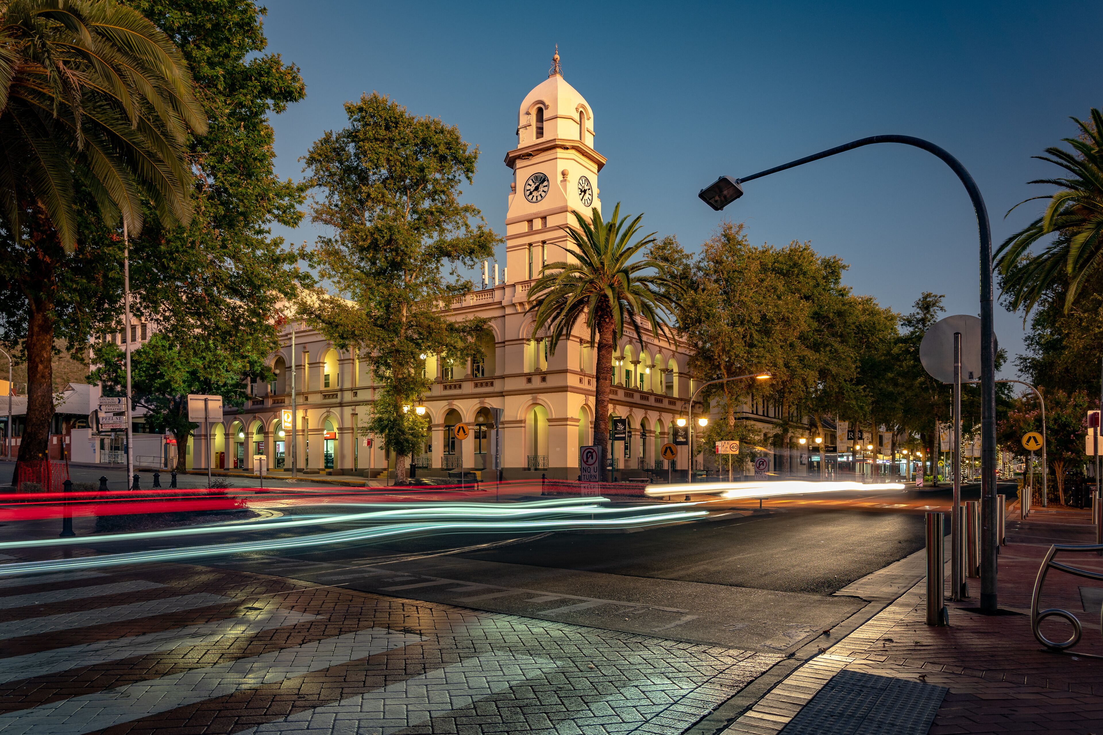 Un edificio histórico con una torre de reloj, palmeras y una calle bien iluminada al anochecer.
