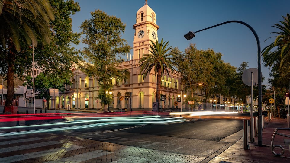 Un edificio histórico con una torre de reloj, palmeras y una calle bien iluminada al anochecer.