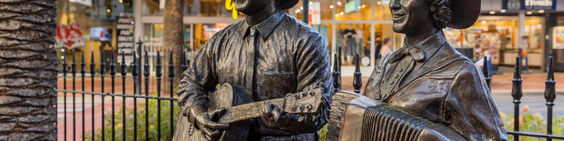 Duas estátuas de bronze retratam homens tocando guitarra e acordeão, com o cenário de uma área comercial com palmeiras e uma cerca.