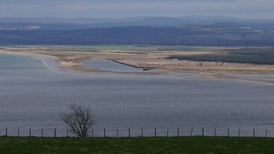A coastal landscape with a sandy beach, a small water body, and distant hills.