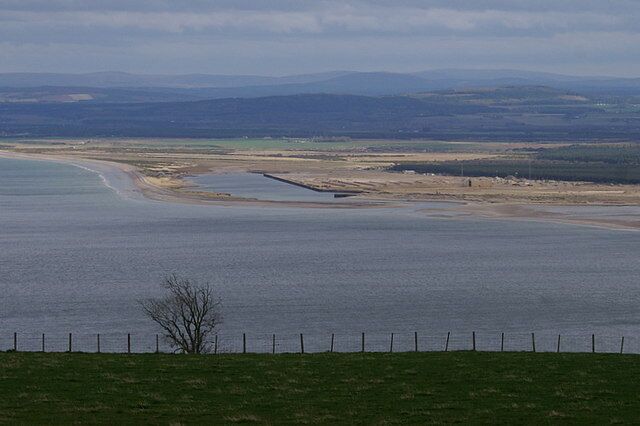 A coastal landscape with a sandy beach, a small water body, and distant hills.