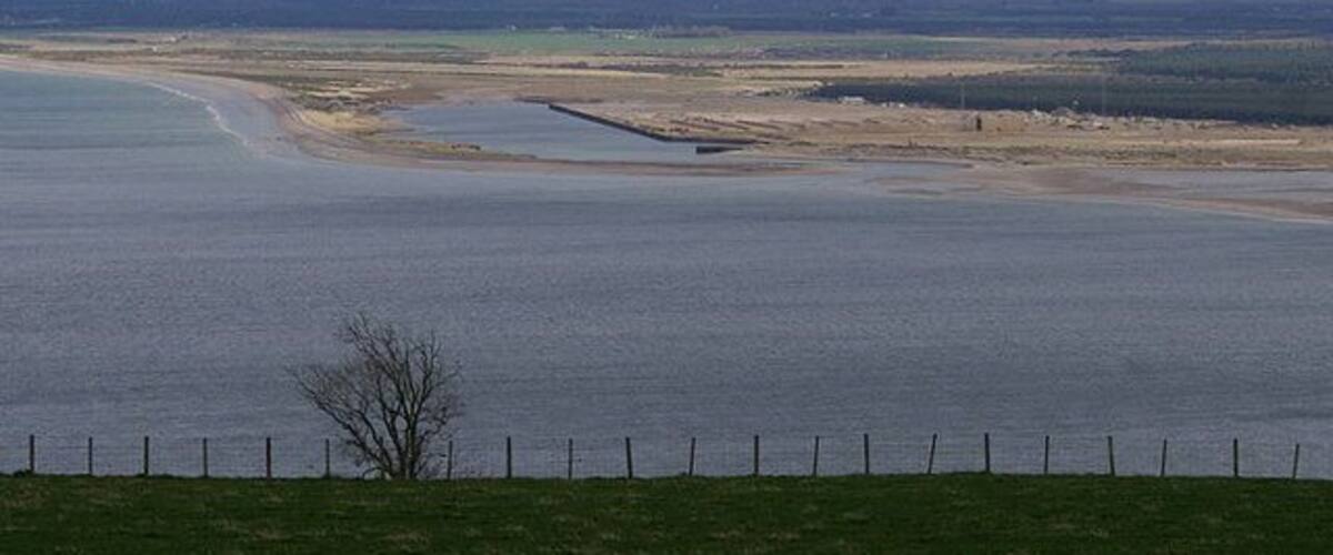 A coastal landscape with a sandy beach, a small water body, and distant hills.