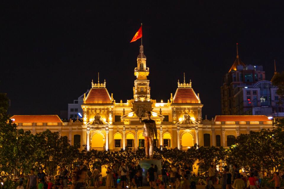 Un edificio histórico y luminoso con una torre central y dos torres más pequeñas, rodeado de árboles y una multitud de personas.